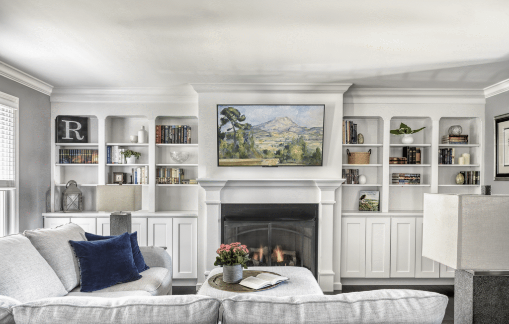 Bright living room with white custom built-in bookshelves flanking a central fireplace with a framed TV displaying landscape art.