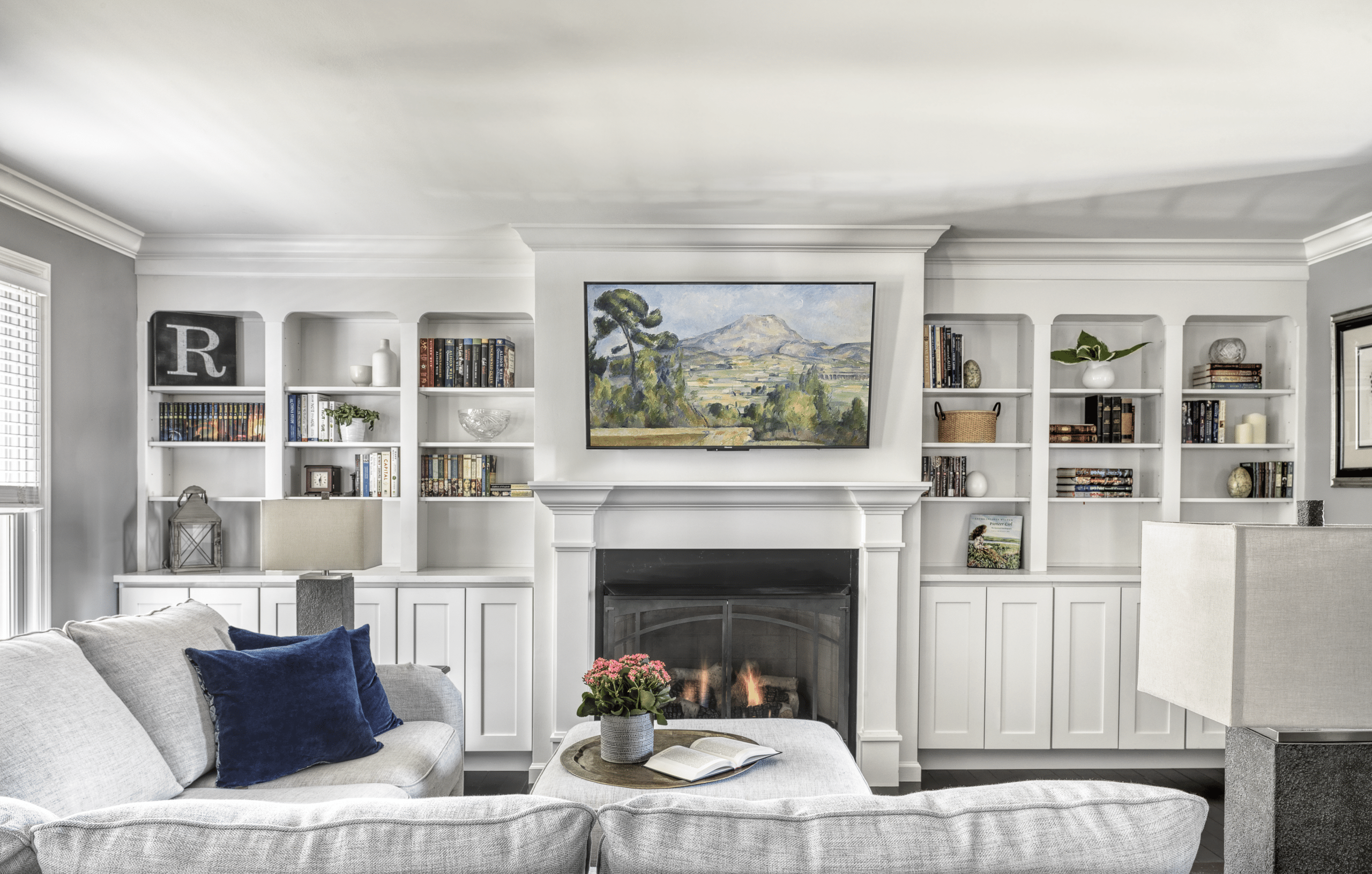 Bright living room with white custom built-in bookshelves flanking a central fireplace with a framed TV displaying landscape art.